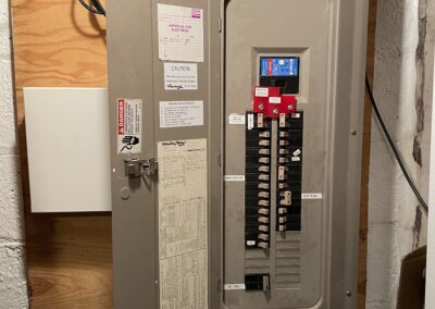 An open electrical panel with multiple labeled circuit breakers mounted on a wooden board in a basement.