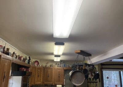 A kitchen with wooden cabinets, fluorescent lights on the ceiling, hanging pots and pans, and various kitchen appliances visible.