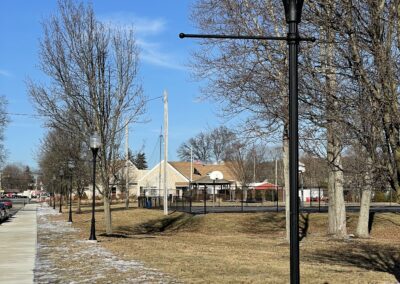 A row of street lamps lines a sidewalk beside a grassy area with sparse snow patches. Bare trees stand nearby under a clear blue sky. A playground and tents are visible in the background.