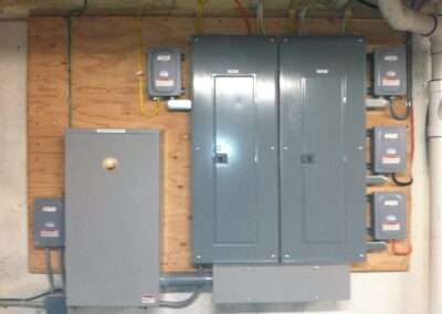 Electrical panel setup with multiple gray circuit boxes mounted on a wooden board against a wall. Various colored wires are connected to the units.